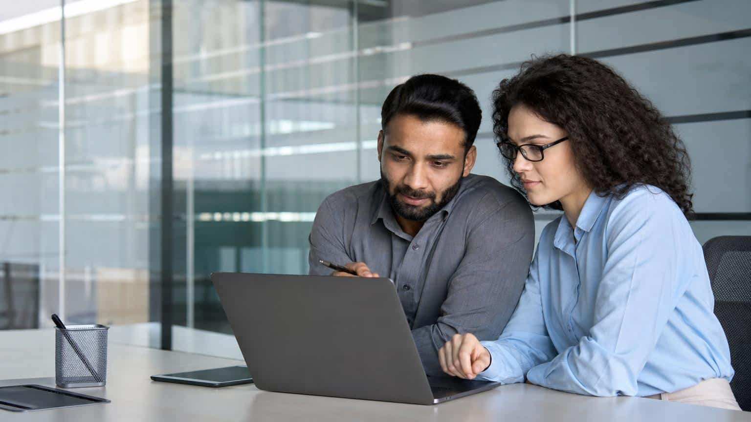 Two colleagues in professional attire collaborate while looking at a laptop screen in a modern office with glass partitions.