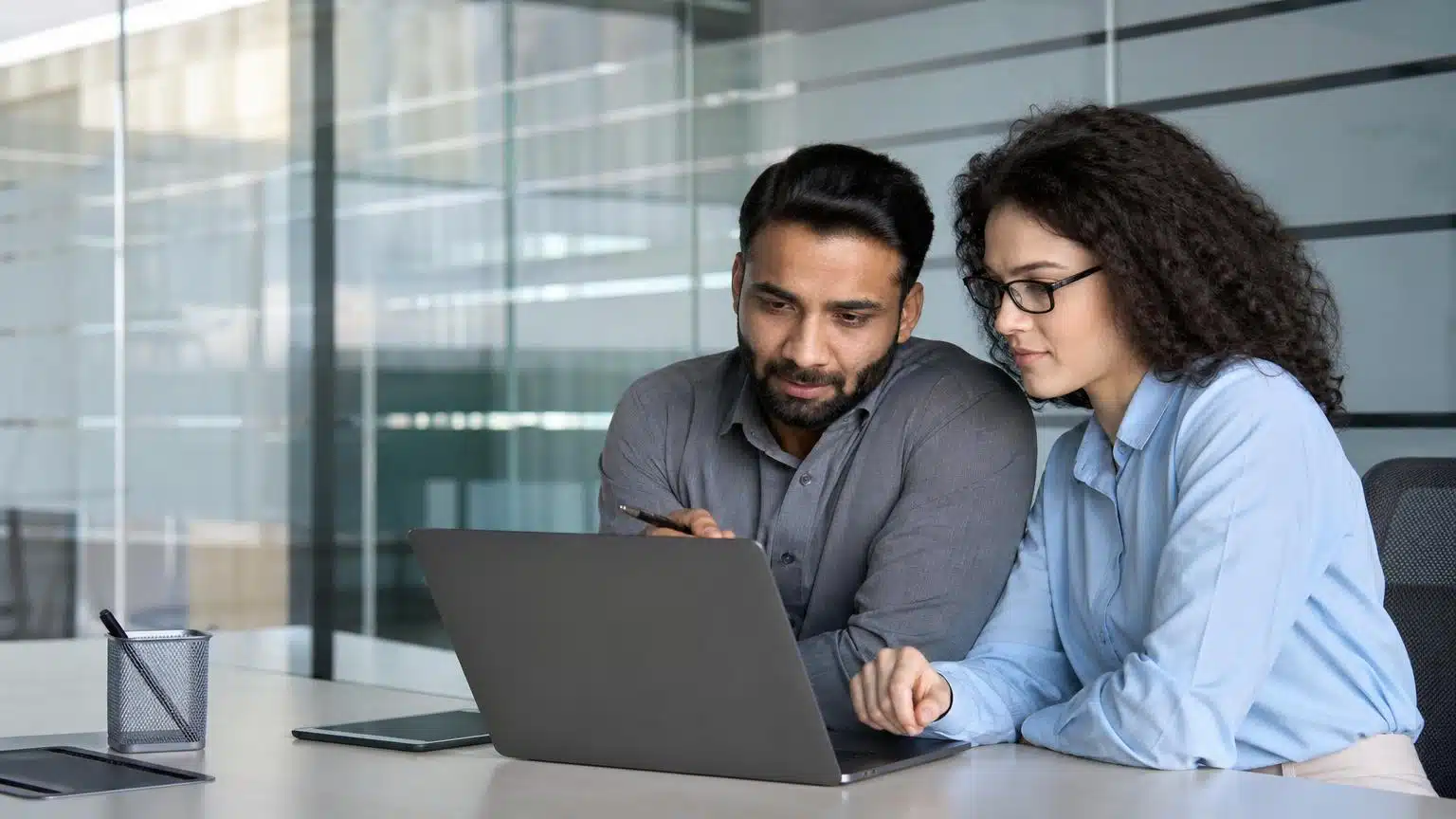 Two colleagues in professional attire collaborate while looking at a laptop screen in a modern office with glass partitions.